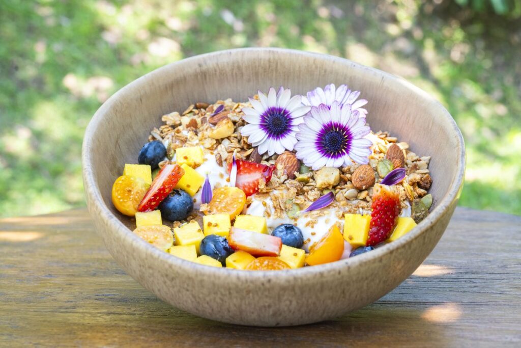 A stoneware bowl of granola with mango, strawberry, blueberries and nuts, finished with purple edible flowers, photographed on the grass