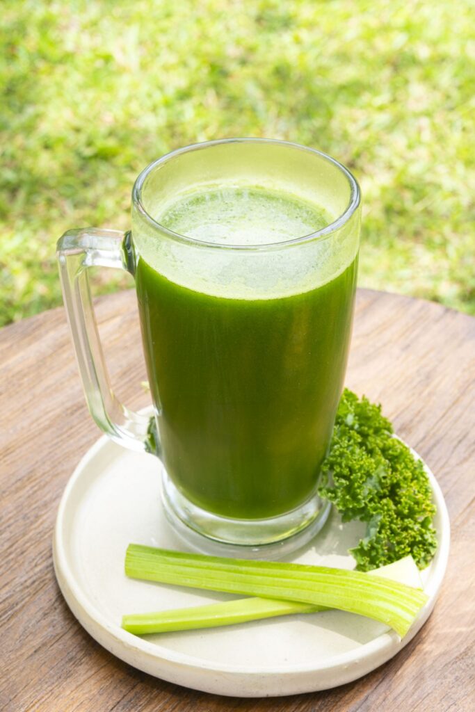 A tall glass of green juice on a saucer, with celery sticks and parsley laid beside it