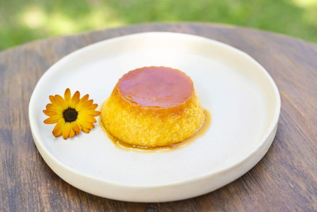 A single flan in caramel on a white plate, with a yellow edible flower alongside