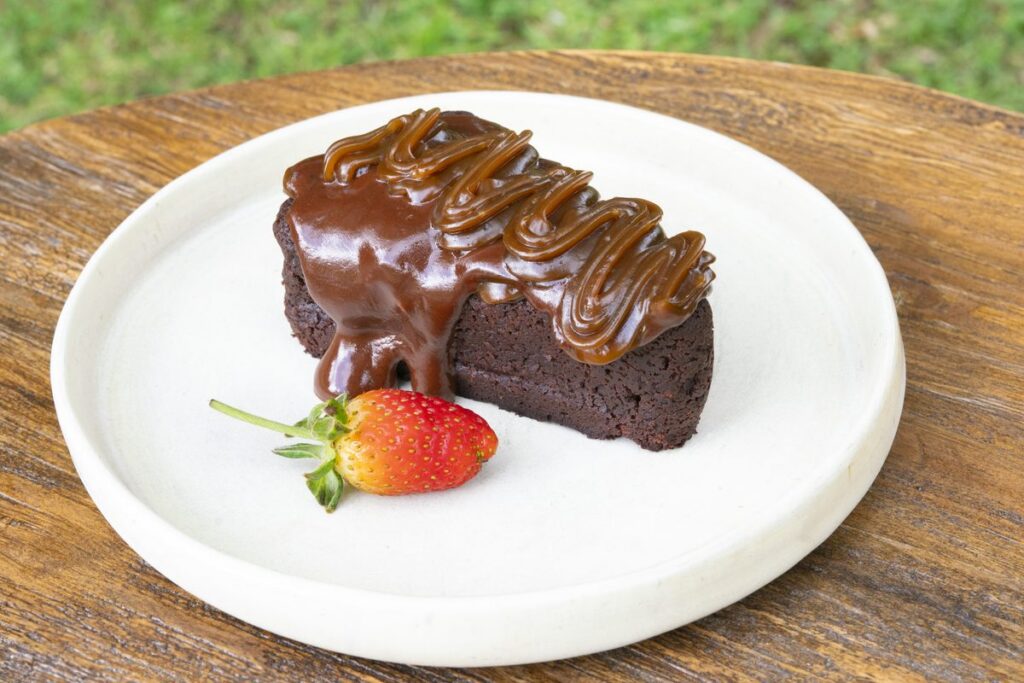 A slice of dark chocolate cake under a chocolate drizzle, with a fresh strawberry beside it on a white plate