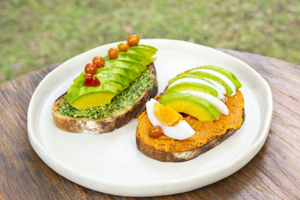 Two open toasts on a white plate — one with pesto and fanned avocado, one with a red pepper spread, avocado and egg