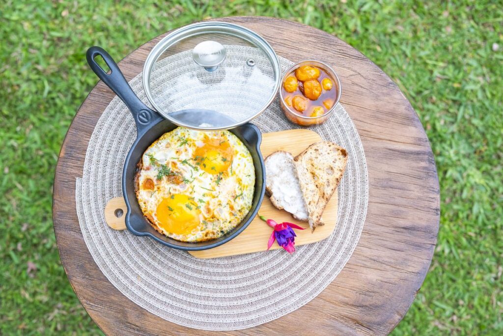 Baked eggs in a small cast-iron skillet with a glass lid, served with bread and a side of stewed tomato, on a woven mat