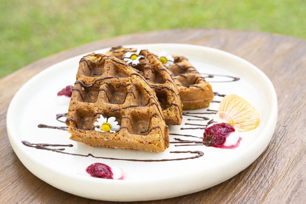 Waffles fanned across a white plate with chocolate sauce, raspberries, cream and a white edible flower