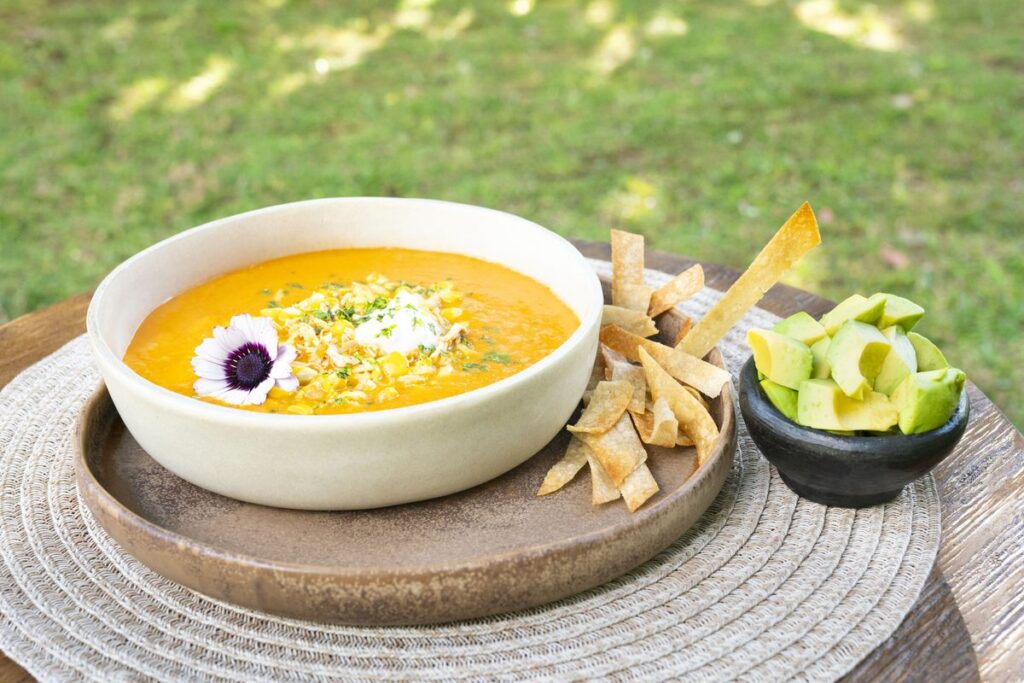 A bowl of golden squash soup with herbs and an edible flower, served with crisp strips and a small dish of avocado