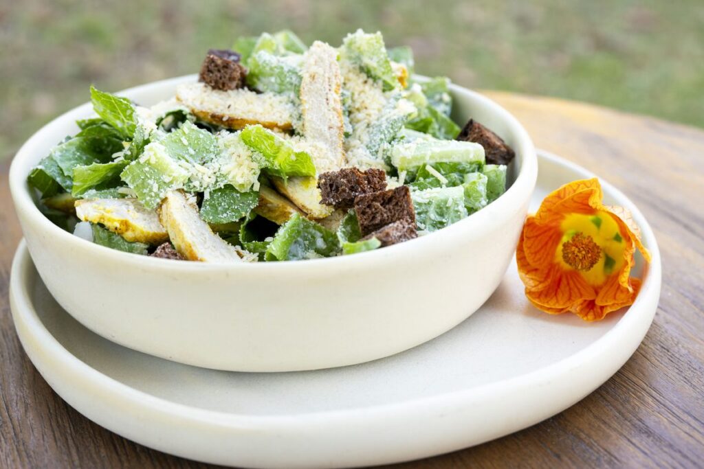 A deep bowl of green salad with dark rye croutons and shaved cheese, with an orange edible flower beside it