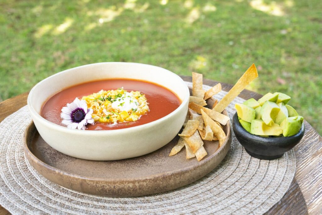 A bowl of tomato soup with herbs and an edible flower, served with crisp strips and a small dish of avocado