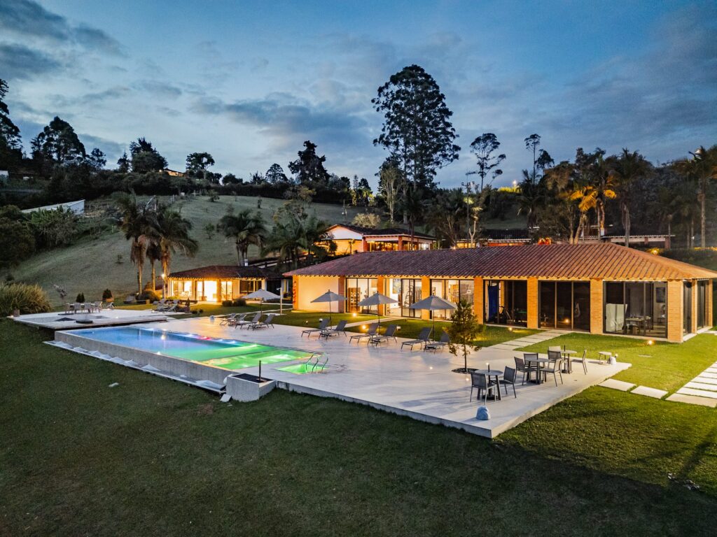 The main house and lap pool at dusk, the interiors lit warm, with the treeline and evening sky behind
