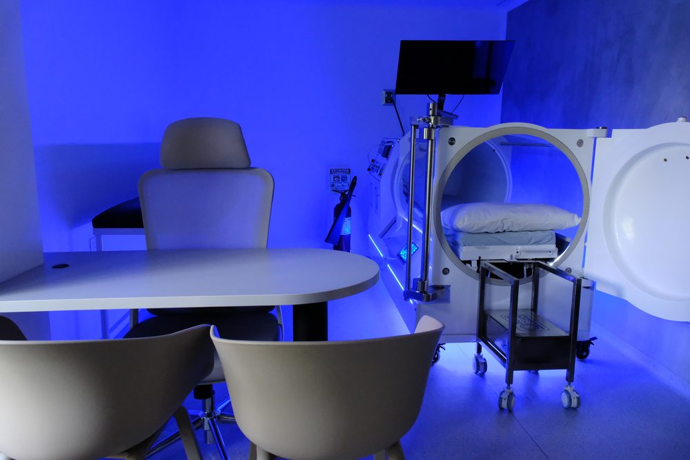 The hyperbaric oxygen chamber standing open in a treatment room lit deep blue, with a consultation table and chairs alongside
