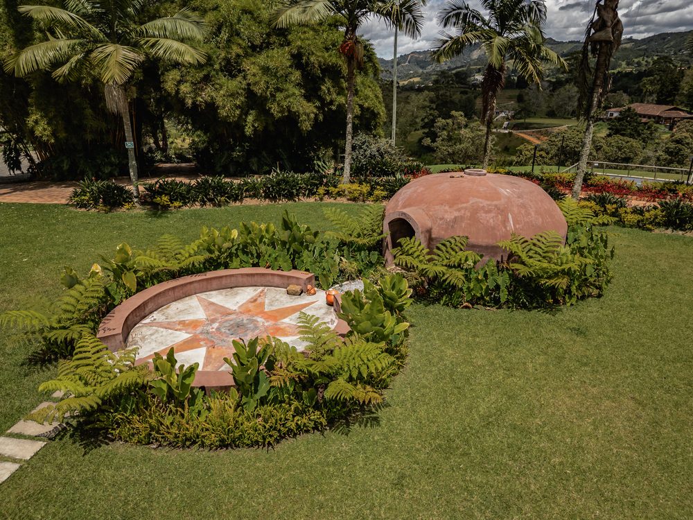 The temazcal dome among ferns and palms, with a tiled sun motif set into the ground and the valley beyond