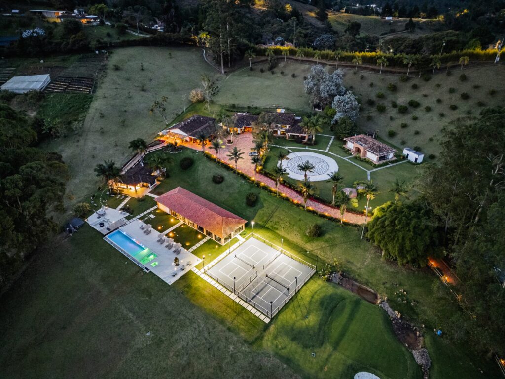 Aerial view of the property at dusk, the buildings lit warm against the lawns, with the pool and courts below