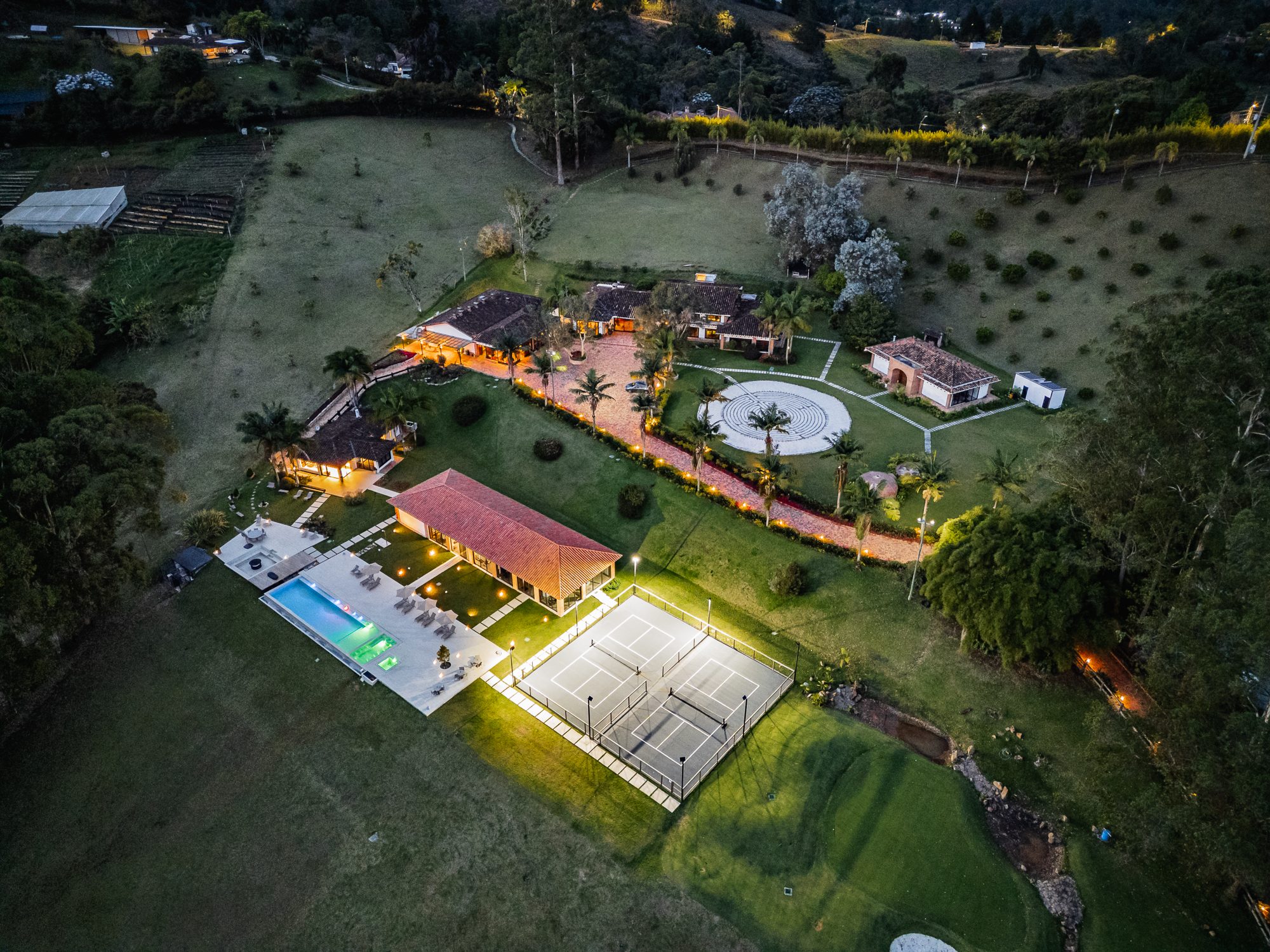Aerial view of the property at dusk, the buildings lit warm against the lawns, with the pool and courts below