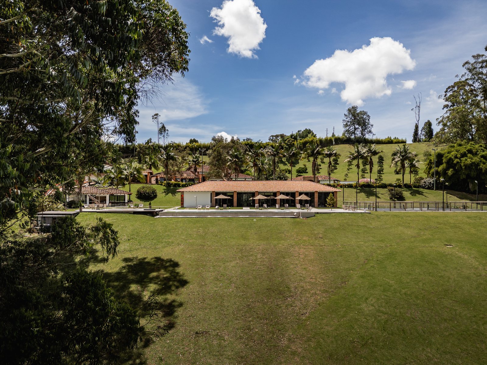 A wide open lawn running down to the long open-sided pavilion, framed by trees under a bright sky