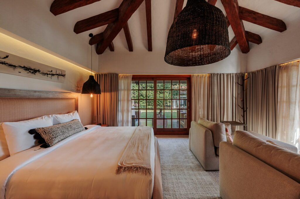 A guest room under exposed timber beams, with a large bed, a woven pendant lamp and doors opening to the garden